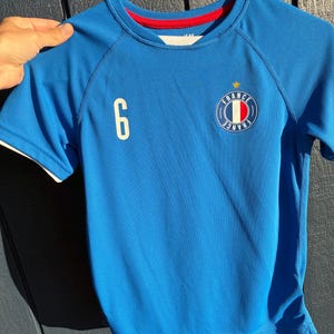 May include: A blue short-sleeved athletic jersey featuring a white number 6 on the right chest and a circular emblem on the left chest with the text FRANCE, a central tricolor stripe, and a small yellow star above the circle. The collar has red trim.