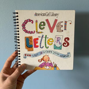 May include: A spiral-bound book titled American Girl Library Clever Letters: Fun ways to wiggle your words. The cover features colorful, stylized lettering, a cartoon illustration of a person writing at a desk, and various decorative elements.
