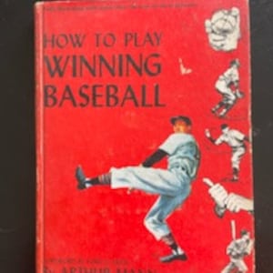 Puede incluir: Un libro rojo de tapa dura titulado "How to Play Winning Baseball" de Arthur Mann. La portada presenta a un jugador de béisbol con un uniforme azul pateando su pierna para lanzar una pelota.