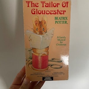 May include: A VHS video cassette featuring a family musical for Christmas based on the book "The Tailor of Gloucester" by Beatrix Potter. The cover features a cartoon mouse holding a book and a spool of thread.