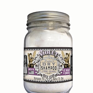 May include: A glass jar with a silver lid containing a white powder. The label reads "Violet The Apawthecary Fairy Dry Shampoo Lavender Chamomile Scent" and "Net Weight 12 Ounces".