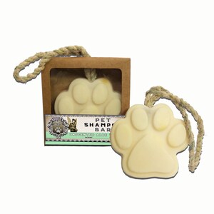 May include: A light yellow, paw-shaped pet shampoo bar with a rope loop, displayed next to its cardboard box. The box features a label with the text "Pet Shampoo Bar" and "Unscented Aloe". The bar is designed for pet grooming.