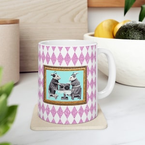 May include: White ceramic mug with a pink and white diamond pattern. The mug features a framed image of two anthropomorphic animals with headphones, operating a vintage radio on a light blue background. The mug sits on a beige coaster.