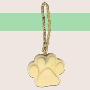 May include: A light yellow paw print-shaped soap with a woven rope loop for hanging. The soap has a smooth surface and is suspended against a background with a green stripe.