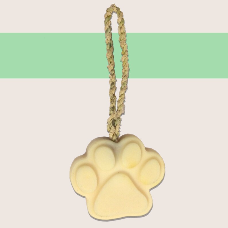 May include: A light yellow paw print-shaped soap with a woven rope loop for hanging. The soap has a smooth surface and is suspended against a background with a green stripe.