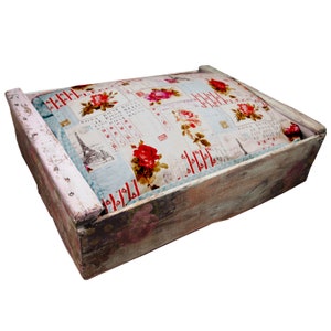 Puede incluir: Una cama de perro de madera con un acabado de pintura blanca desgastada. La cama tiene un cojín con un patrón floral con rosas rojas y las palabras "Rosa Rosa Rosa" repetidas.