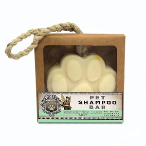 May include: A pet shampoo bar shaped like a paw print, in a brown cardboard box with a clear window. The label reads "Unscented Aloe" and "Net Weight 5 ounces". A rope handle is attached to the box.