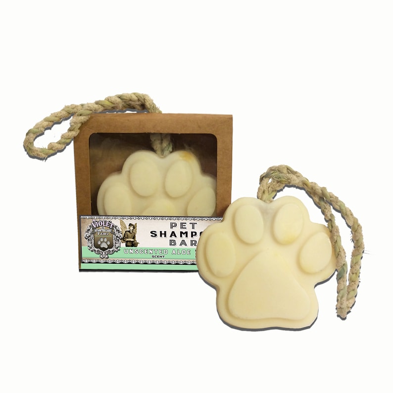 May include: A light yellow, paw-shaped pet shampoo bar with a rope loop, displayed next to its cardboard box. The box features a label with the text "Pet Shampoo Bar" and "Unscented Aloe". The bar is designed for pet grooming.
