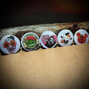 May include: A collection of five round buttons with Halloween and horror-themed designs. The buttons feature images of a skull with flower eyes, a Camp Crystal Lake logo, a heart with the words "Call Me" and a ghost face, a Hello Kitty witch, and a jack-o'-lantern.