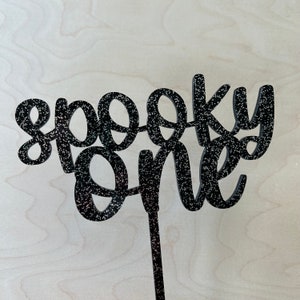 May include: Black glitter cake topper with the words "spooky one" in a whimsical font.