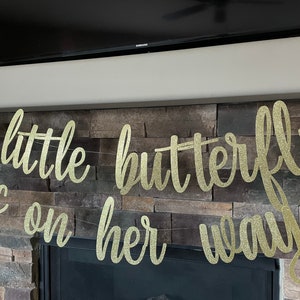 May include: A gold glitter banner with the words "a little butterfly is on her way" in cursive script. The banner is strung across a stone fireplace. A butterfly decoration is visible on the left.
