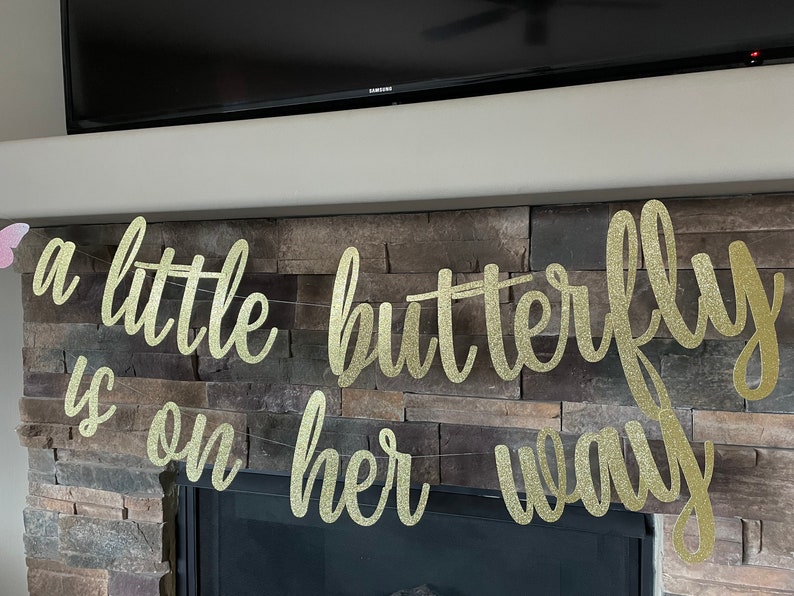 May include: A gold glitter banner with the words "a little butterfly is on her way" in cursive script. The banner is strung across a stone fireplace. A butterfly decoration is visible on the left.