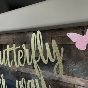 May include: A gold glitter banner with the words "the butterfly" and "on her way" in a cursive font. A pink glitter butterfly is strung on the same line. The banner is hanging in front of a stone wall.