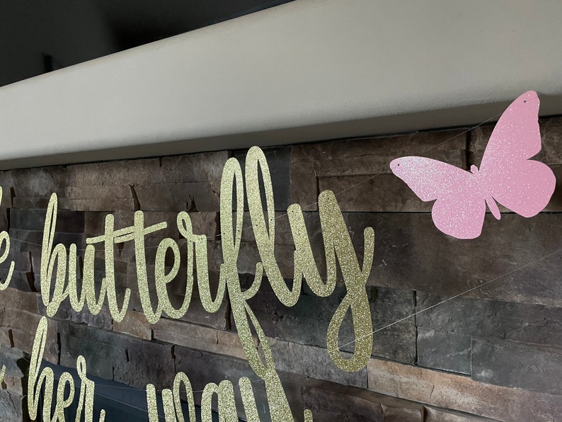 May include: A gold glitter banner with the words "the butterfly" and "on her way" in a cursive font. A pink glitter butterfly is strung on the same line. The banner is hanging in front of a stone wall.
