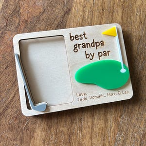 May include: Wooden photo frame with a golf theme. The frame has a cutout of a golf club and a green golf course with a flag. The text reads "best grandpa by par" and "Love. Jude. Dominic. Max. & Leo".