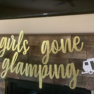 May include: A gold glitter banner that reads "girls gone glamping" in cursive script. Two white camper cutouts flank the text. The banner is displayed against a stone fireplace.