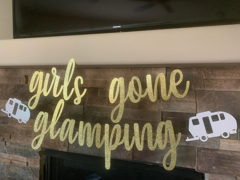 May include: A gold glitter banner that reads "girls gone glamping" in cursive script. Two white camper cutouts flank the text. The banner is displayed against a stone fireplace.