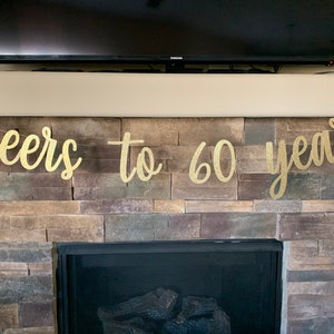 May include: A gold glitter banner that reads "cheers to 60 years" is displayed above a fireplace. The banner is made of a shiny material and is strung across the top of the fireplace. The fireplace is made of stone.