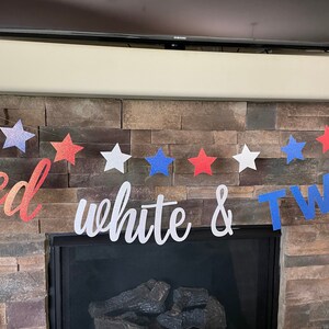 May include: A festive banner with red, white, and blue star shapes and the words "red white & TWO". The banner is strung across a stone fireplace, perfect for a patriotic or birthday celebration.