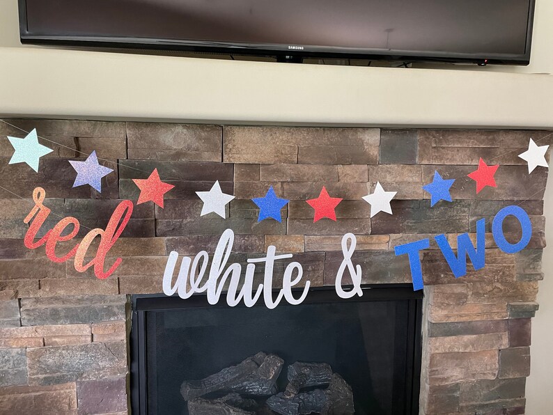 May include: A festive banner with red, white, and blue star shapes and the words "red white & TWO". The banner is strung across a stone fireplace, perfect for a patriotic or birthday celebration.