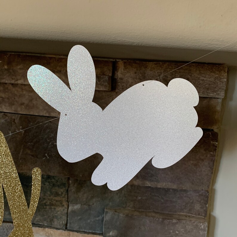 May include: A glittery silver bunny-shaped banner cutout, part of a festive decoration. The bunny is strung on a thin, clear string, ready for hanging. The background is a textured, dark stone wall.