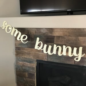 May include: A glittery gold banner that reads "some bunny" in a cursive font. The banner is strung across a stone fireplace, with a black television mounted above.