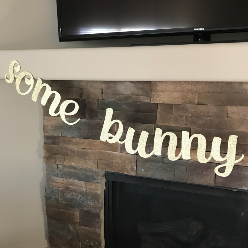 May include: A glittery gold banner that reads "some bunny" in a cursive font. The banner is strung across a stone fireplace, with a black television mounted above.