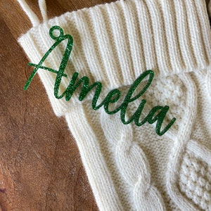 May include: White knitted stocking with a green glitter name tag that says "Amelia".