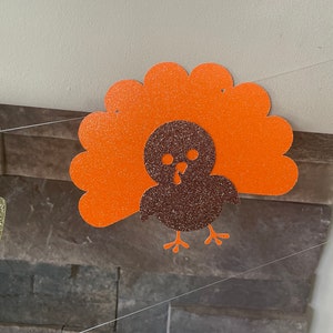 May include: A glittery orange and brown turkey decoration, likely for Thanksgiving. The turkey has an orange fan-shaped tail and a brown body. The decoration is hanging from a string, with a stone wall in the background.
