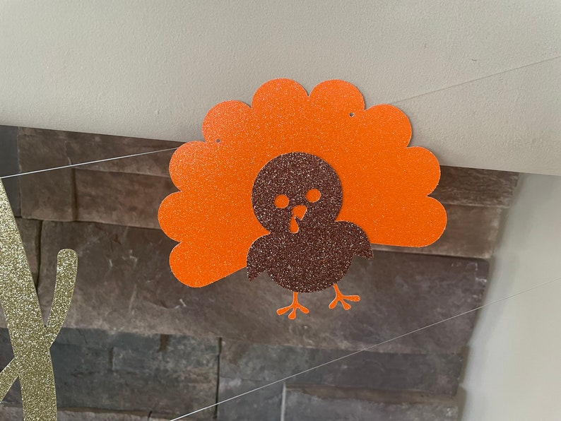 May include: A glittery orange and brown turkey decoration, likely for Thanksgiving. The turkey has an orange fan-shaped tail and a brown body. The decoration is hanging from a string, with a stone wall in the background.
