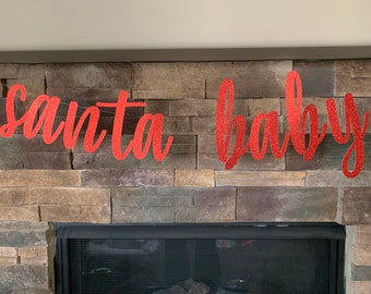 Peut inclure: Une bannière rouge à paillettes avec l'inscription "santa baby" en écriture cursive. La bannière est suspendue sur un mur de pierre de couleur neutre, au-dessus d'une cheminée.