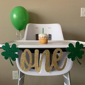 May include: A white high chair with a green shamrock banner that says "one" in gold glitter. A green balloon and a cupcake with a green candle are on the tray.