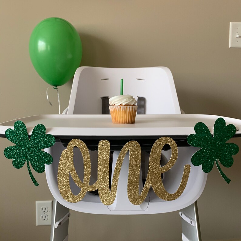May include: A white high chair with a green shamrock banner that says "one" in gold glitter. A green balloon and a cupcake with a green candle are on the tray.