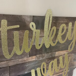 May include: A gold glitter banner with the words "turkey" and "eay" in a cursive font. The banner is strung across a stone fireplace. An orange decoration is visible on the right side.