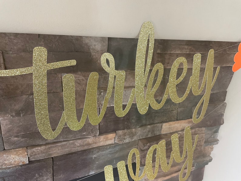 May include: A gold glitter banner with the words "turkey" and "eay" in a cursive font. The banner is strung across a stone fireplace. An orange decoration is visible on the right side.