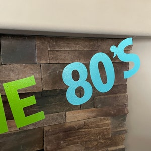May include: A glittery banner with the text "THE 80'S" in lime green and light blue. The banner is strung across a stone wall, creating a festive and retro party decoration.