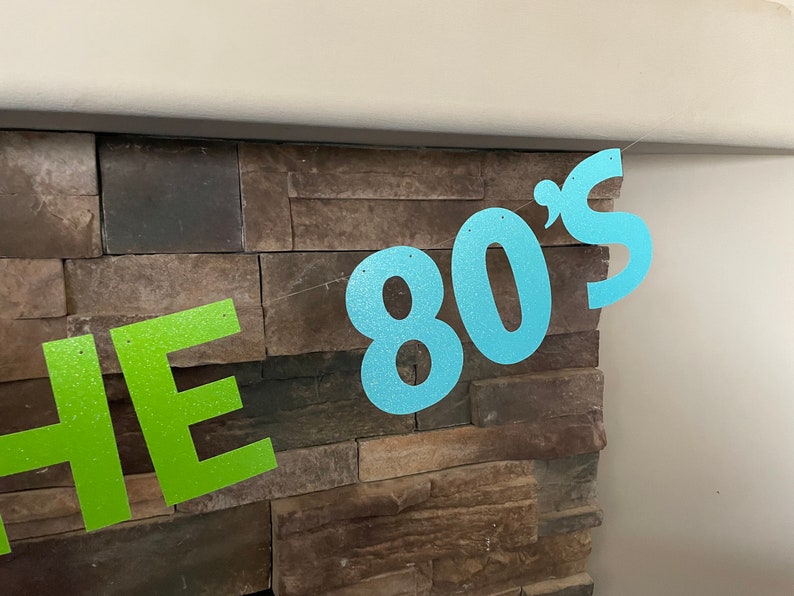May include: A glittery banner with the text "THE 80'S" in lime green and light blue. The banner is strung across a stone wall, creating a festive and retro party decoration.