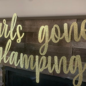 May include: A gold glitter banner that reads "girls gone glamping" in a cursive font. The banner is strung across a stone fireplace. White camper silhouettes are on each end.