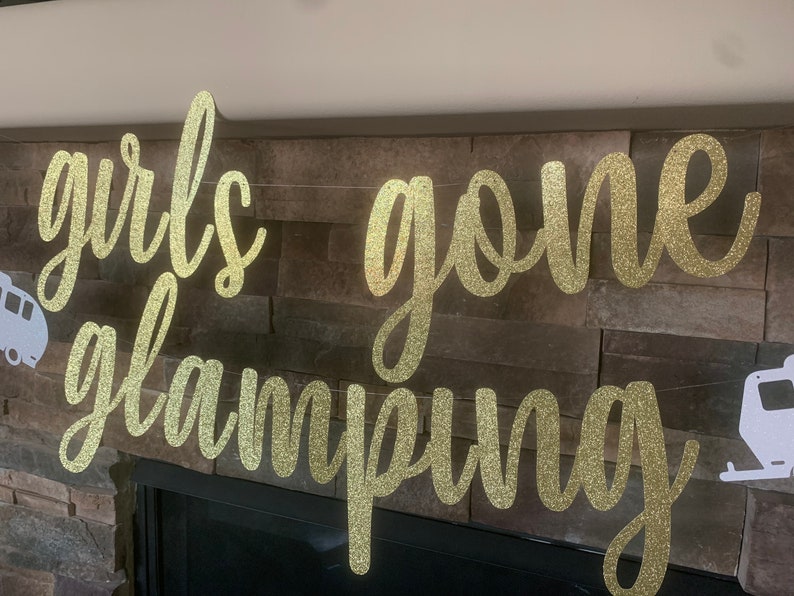 May include: A gold glitter banner that reads "girls gone glamping" in a cursive font. The banner is strung across a stone fireplace. White camper silhouettes are on each end.