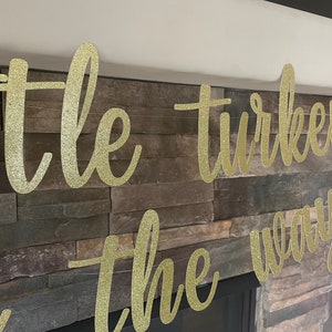 May include: A gold glitter banner that reads "little turkey on the way" in cursive script. The banner is displayed against a stone fireplace. A small turkey decoration is visible on the right.