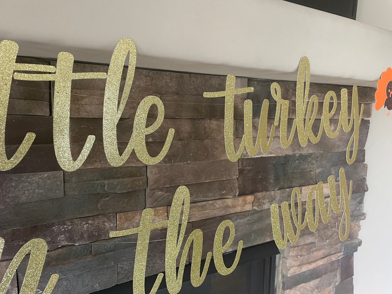 May include: A gold glitter banner that reads "little turkey on the way" in cursive script. The banner is displayed against a stone fireplace. A small turkey decoration is visible on the right.