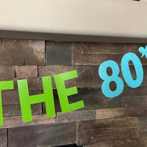 May include: A banner with the text "THE 80's" in glittery green and blue letters. The banner is strung across a stone wall, creating a retro party decoration.