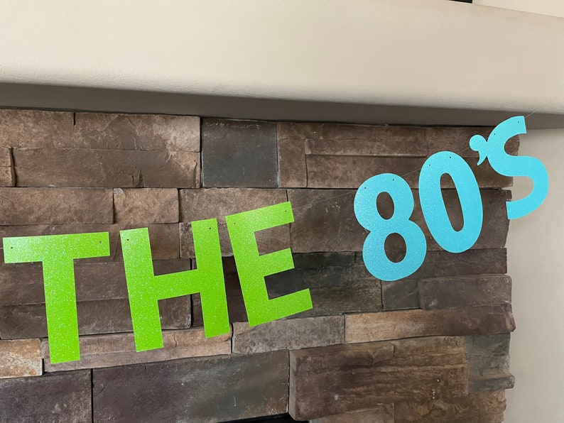 May include: A banner with the text "THE 80's" in glittery green and blue letters. The banner is strung across a stone wall, creating a retro party decoration.
