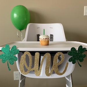 May include: A white high chair with a green shamrock banner that says "one" in gold glitter. There is a green balloon and a cupcake with a single candle on the tray.