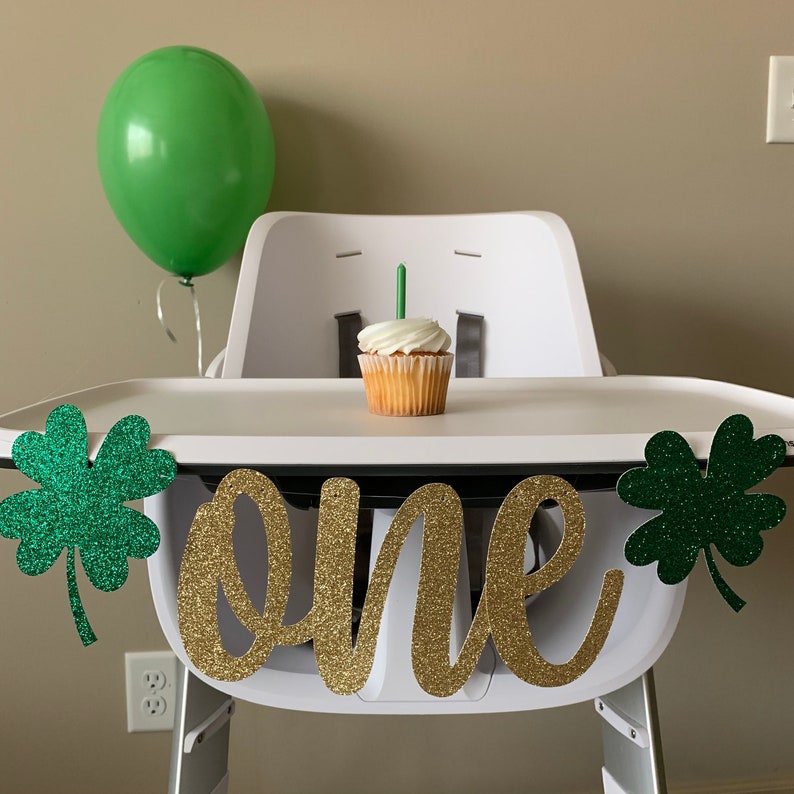 May include: A white high chair with a green shamrock banner that says "one" in gold glitter. There is a green balloon and a cupcake with a single candle on the tray.