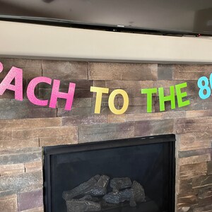 May include: A colorful banner with the text "BACH TO THE 80'S" in glittery, pastel-colored letters. The banner is strung across a stone fireplace, suggesting a party or celebration with an 80s theme.