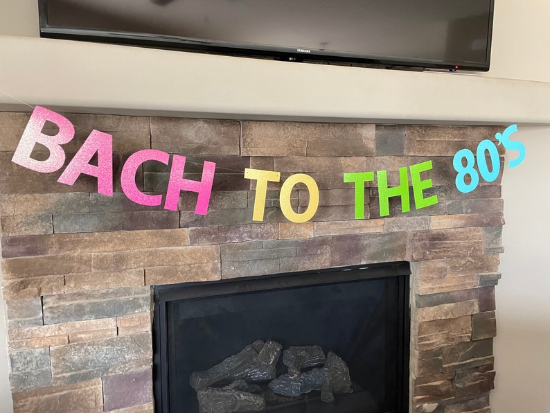 May include: A colorful banner with the text "BACH TO THE 80'S" in glittery, pastel-colored letters. The banner is strung across a stone fireplace, suggesting a party or celebration with an 80s theme.