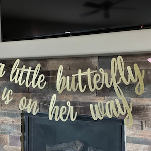 May include: A gold glitter banner with the phrase "a little butterfly is on her way" in cursive script. Two pink butterfly cutouts flank the banner. The banner is strung across a stone fireplace.