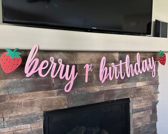 May include: A pink glitter banner that reads "berry 1st birthday" with strawberry accents. The banner is displayed above a fireplace with a stone facade. The strawberries have green glitter tops.