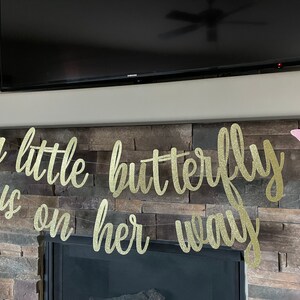 May include: A gold glitter banner with the phrase "a little butterfly is on her way" in cursive script. Pink butterfly cutouts are attached to the banner and placed on either side. The banner is strung across a stone fireplace.